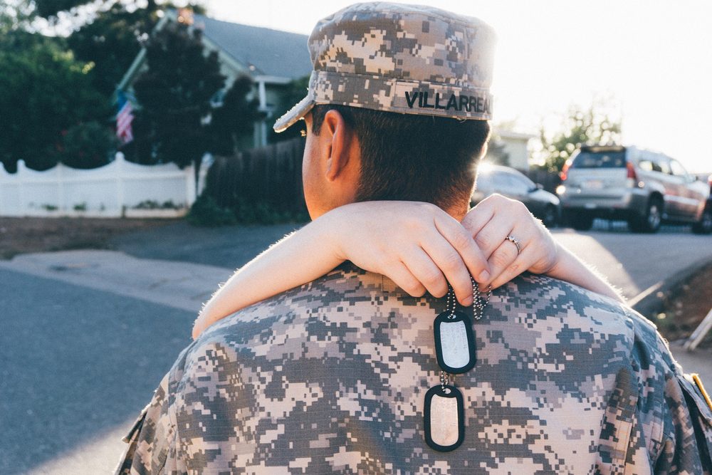 A service member in uniform embraced by a loved one, American flag in the background